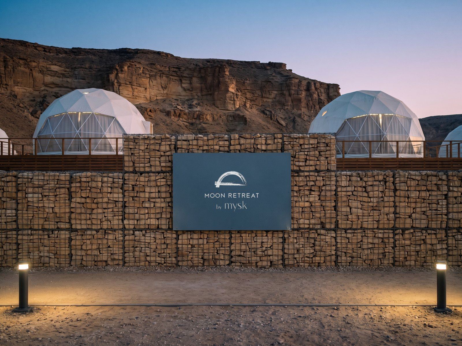 A render: the lockup in white on a slate panel in a gabion wall at blue hour