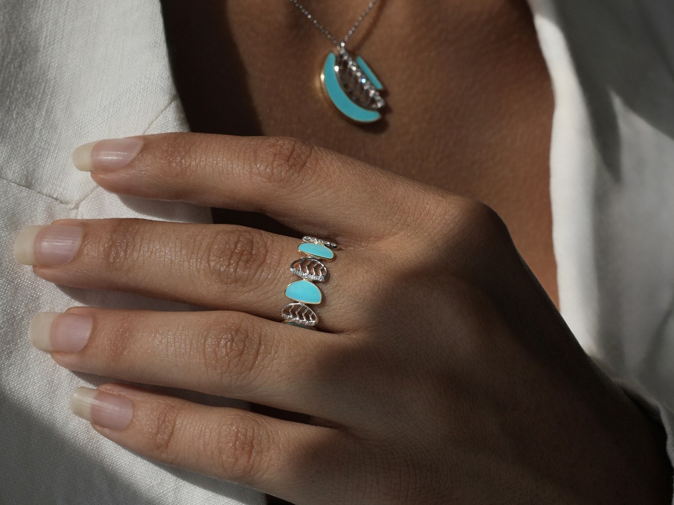 A hand on a white jacket wearing an Amara ring in turquoise enamel, the pendant above